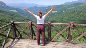 Mujer de espaldas con los brazos abiertos frente a montañas, simbolizando rendirse conscientemente, permitirse suavidad y abrirse al momento presente