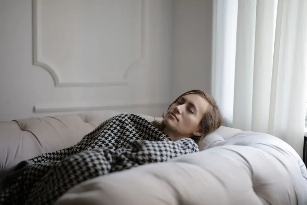 Mujer agotada descansando plácidamente en el sofá tapada con una manta, dándose permiso para sentir su cansancio.